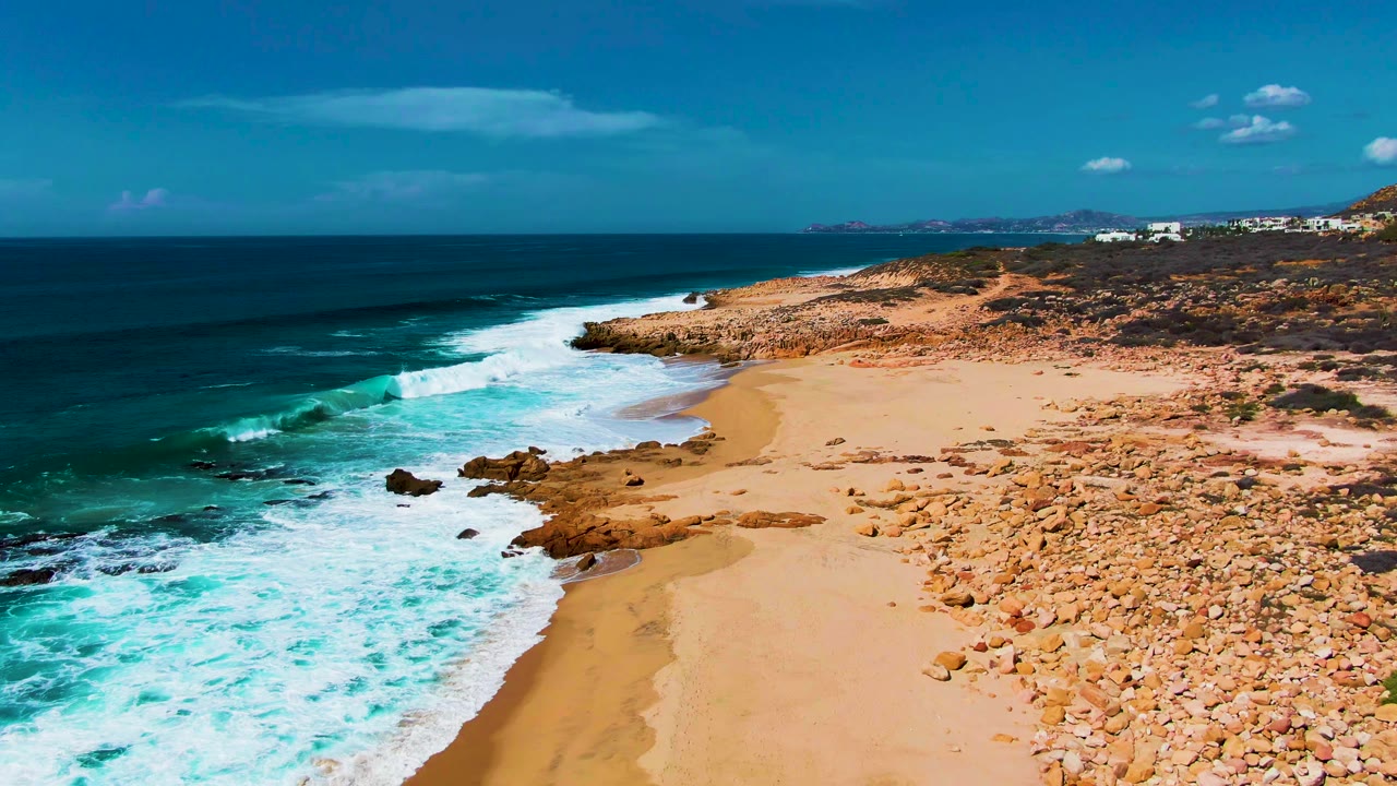 High oblique view of Punta Buzzards surf spreading over shallow turquoise water toward sand and rocks, with low vegetation, far buildings, clear sky, and no visible people.