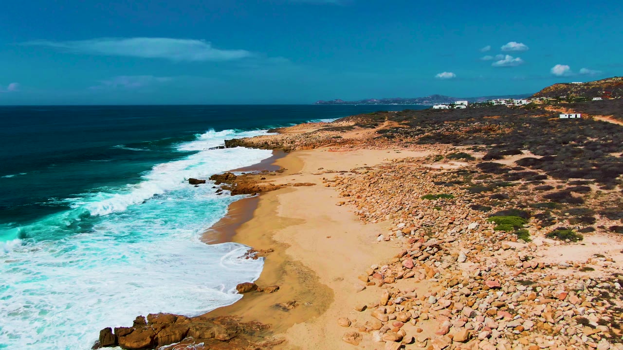Aerial frame of Buzzards Beach in Baja California Sur with turquoise surf washing onto sand, tan rocks along the right side, distant hills, and no visible people.