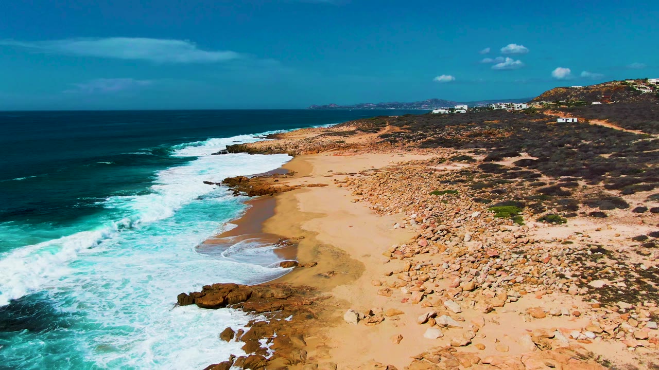 Aerial frame from Buzzards Beach with bright surf breaking around a dark rock outcrop, dry sand curving inland, scattered coastal buildings in the distance, and no visible people.