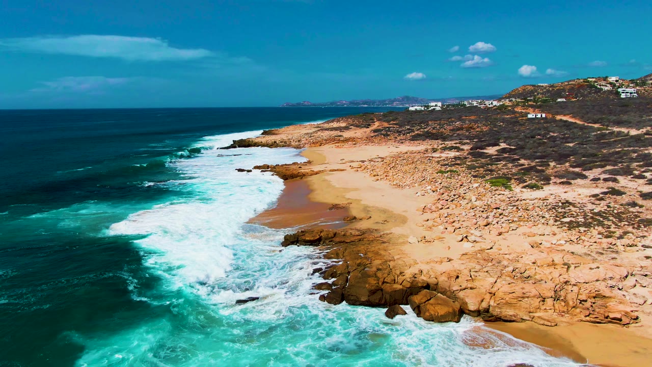 Aerial frame from Punta Buzzards showing turquoise surf breaking against tan rocks and sand, with deep blue ocean at left and no visible people.