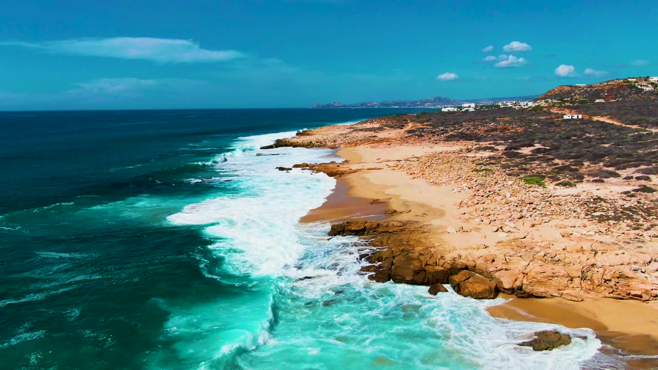 Aerial view at Punta Buzzards with a long white surf line running diagonally along the rocky beach, deep ocean to the left, and no visible people.