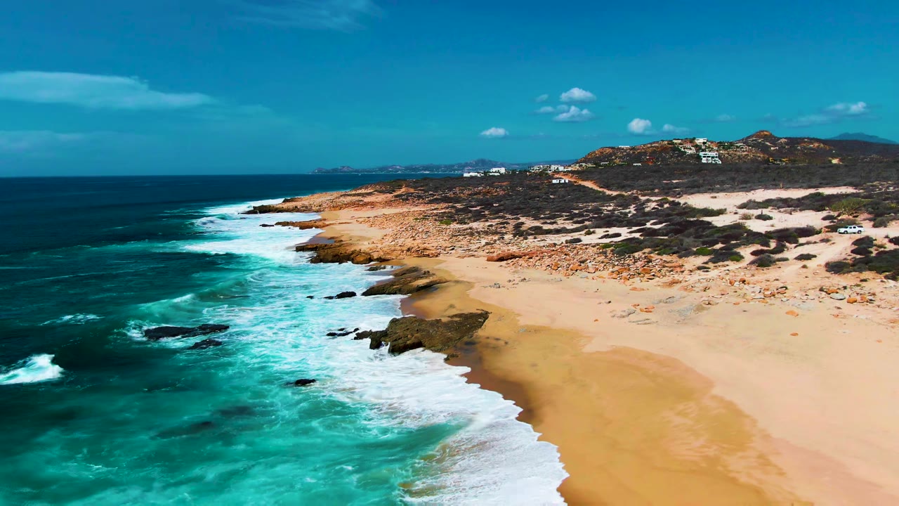 Aerial view of Punta Buzzards coastline with turquoise surf breaking along rocks beside a broad sandy beach, scrubby hills in the background, and no visible people.