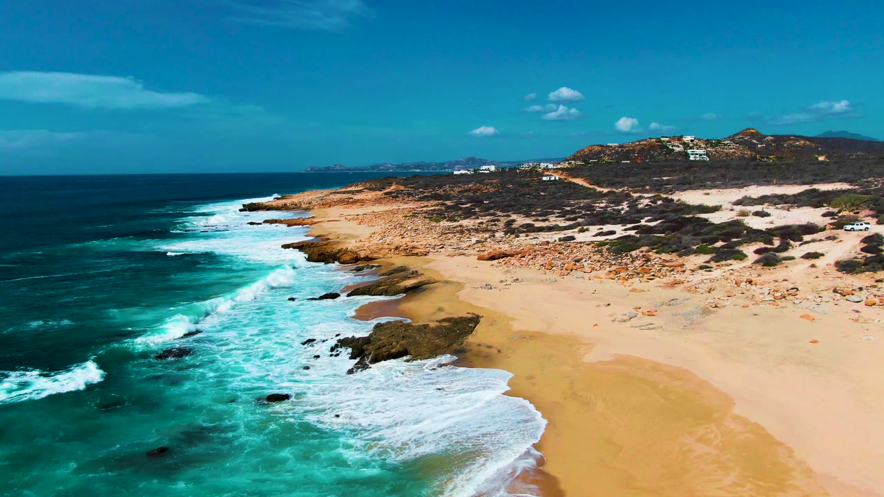 Aerial view over Punta Buzzards as surf curls around several rocky points, leaving wet sand visible near the waterline and open beach to the right, no visible people.