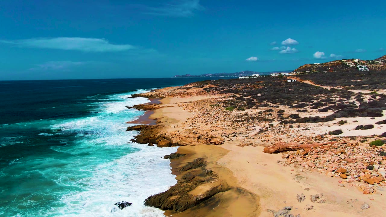 Aerial view at Punta Buzzards, Baja California Sur, with turquoise waves beside a sandy rocky beach, deep blue ocean to the left, low scrub hills in the background, and no visible people.