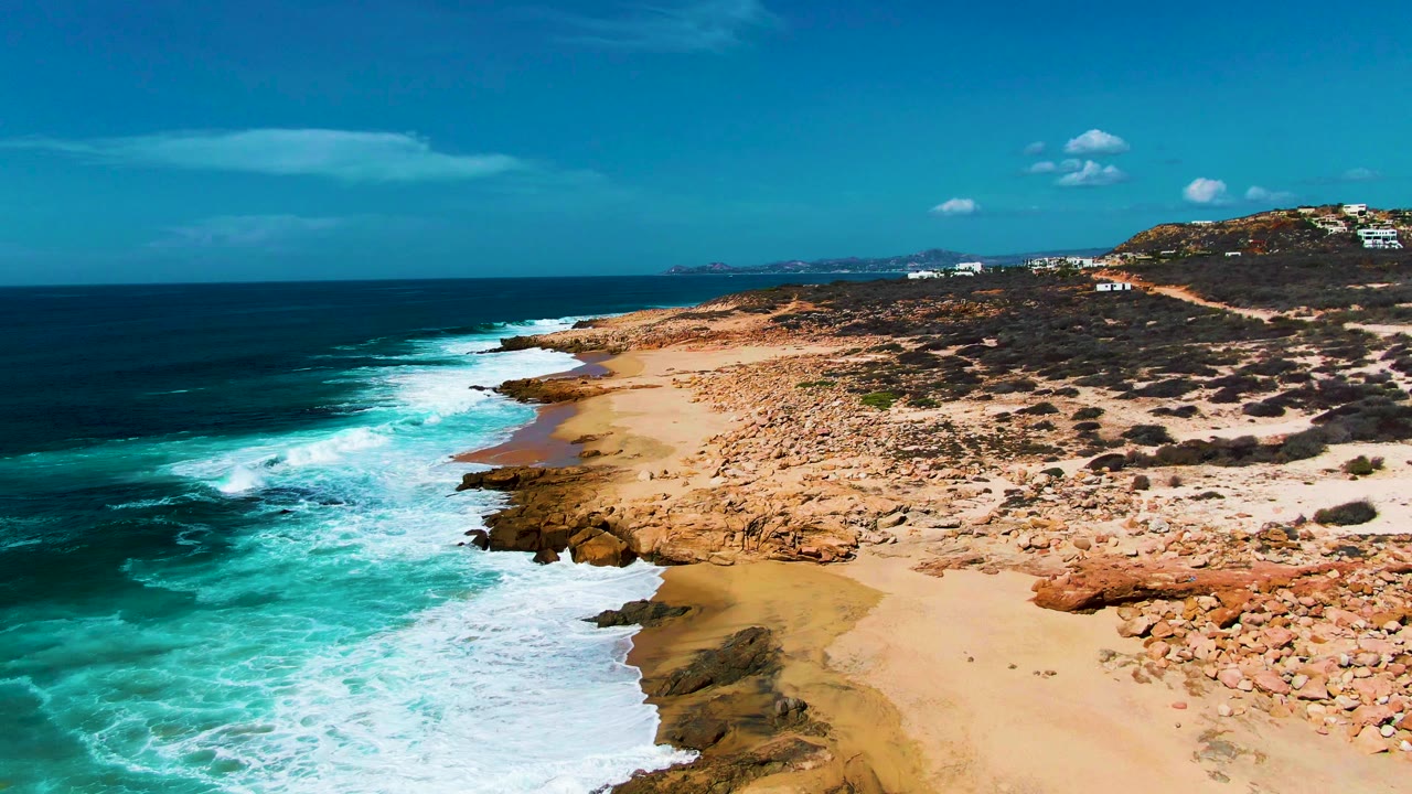 Aerial frame over Punta Buzzards showing whitewater pulling away from the sand, tan rocks lining the cove, dark blue ocean offshore, distant hillside structures, and no visible people.