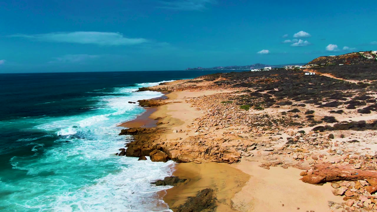 Aerial view of Punta Buzzards with foamy surf spreading across the lower shoreline, a broad tan rock field on the right, deep blue water on the left, and no visible people.