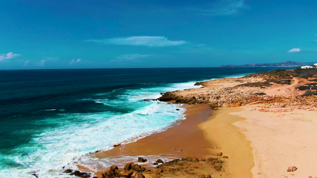 Aerial view at Punta Buzzards, Baja California Sur, showing white surf pushing onto a sandy beach beside rocky shoreline, with no visible people.