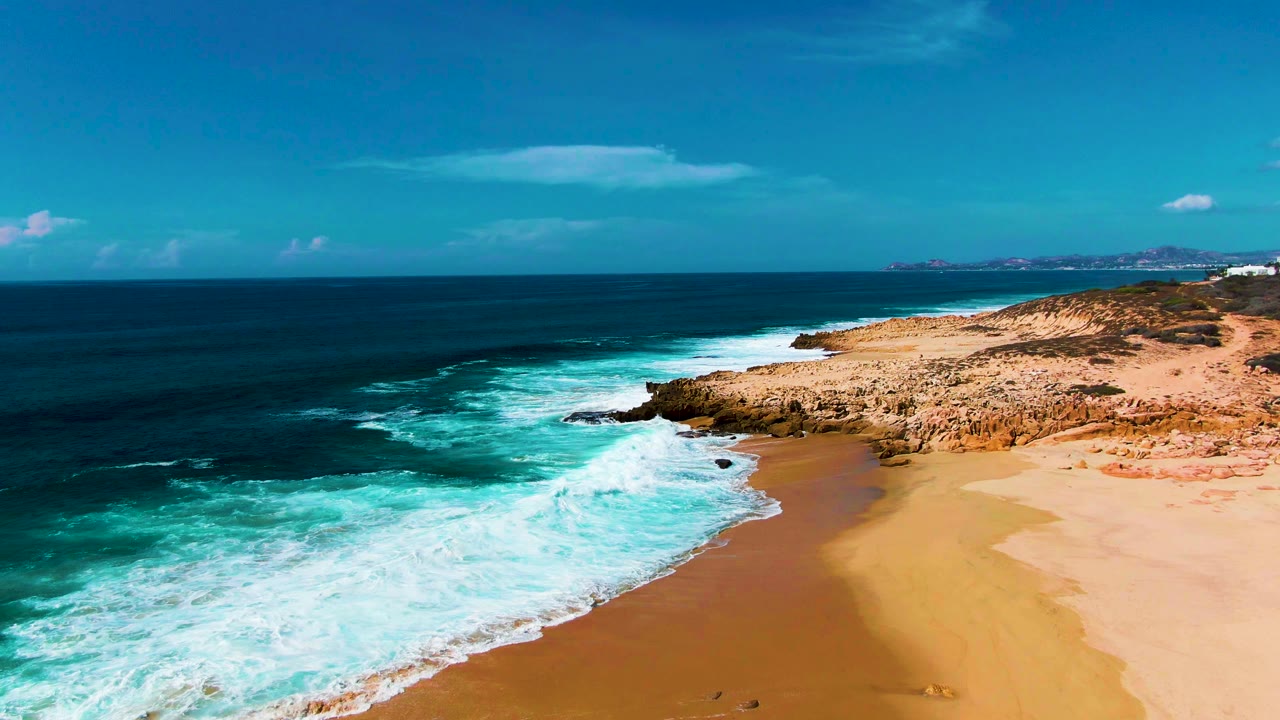 At Punta Buzzards, a band of white foam advances farther up the sandy beach while dark blue water and rocky coast fill the frame, with no visible people.