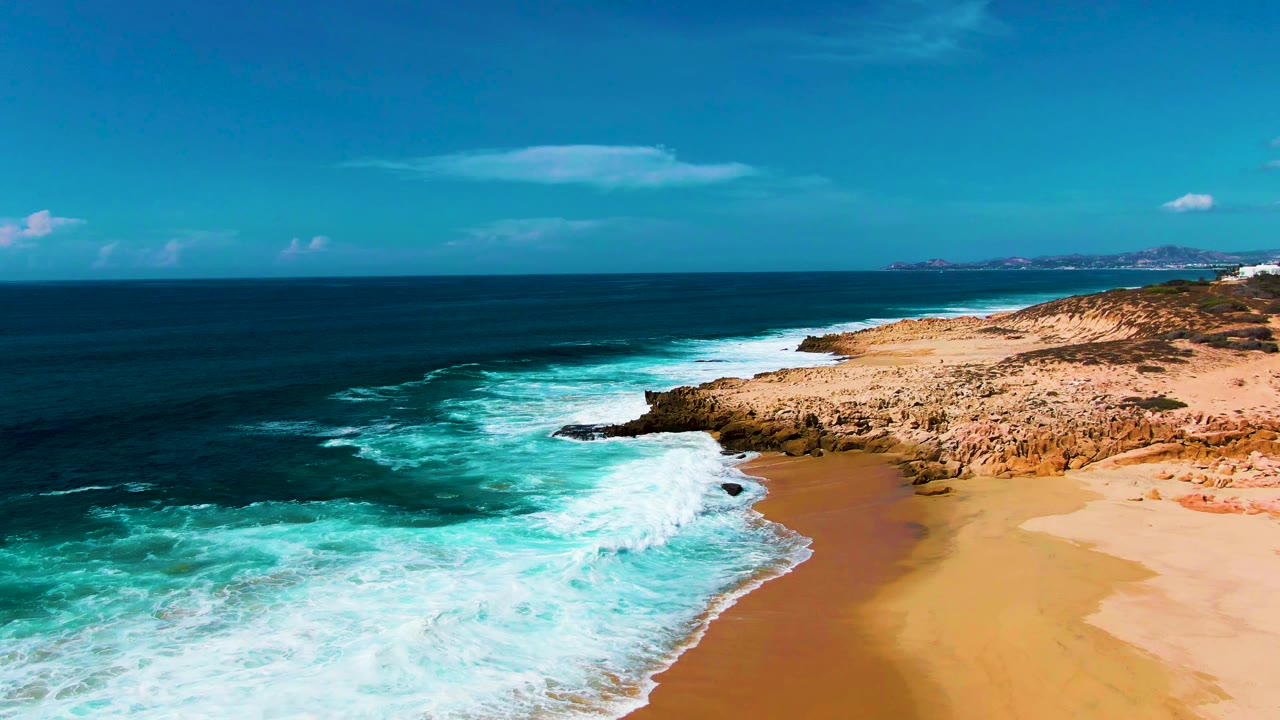 Aerial coastal frame from Punta Buzzards with breaking waves curling toward a rocky point and wet sand, with no visible people on the beach.