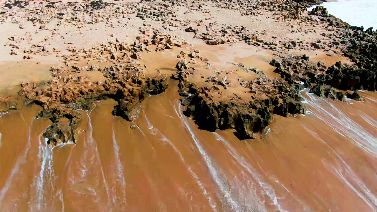 Aerial view of exposed brown sand and dark rocks beside thin surf lines.