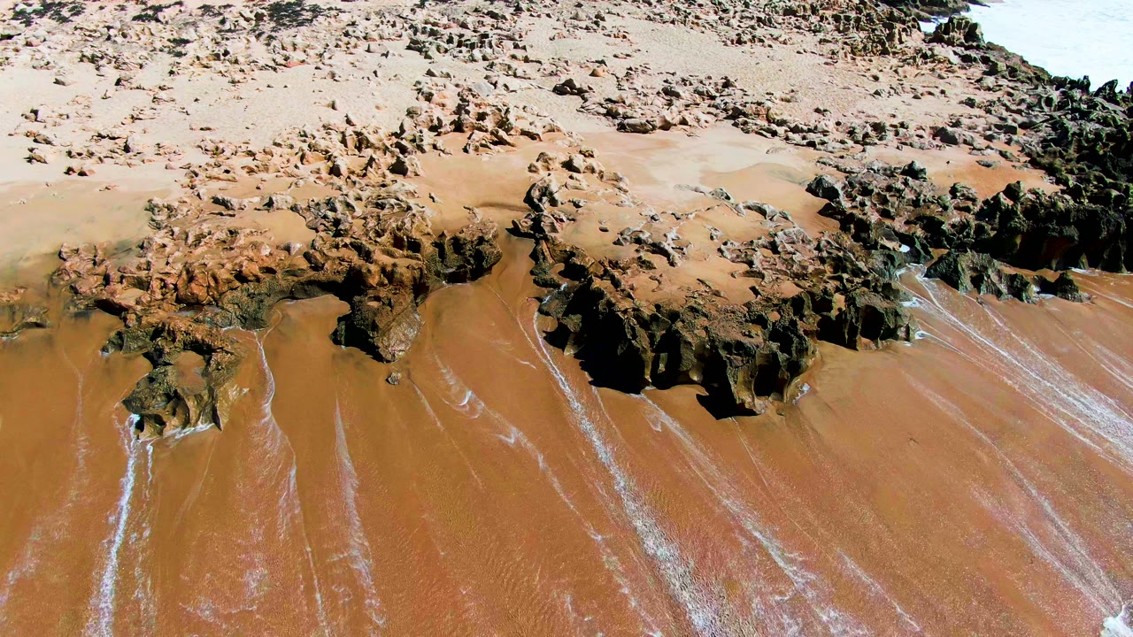 Aerial frame at Punta Buzzards shows the same rocky beach with brown wet sand and narrow foam traces beginning to brighten the lower right edge, with no visible people.