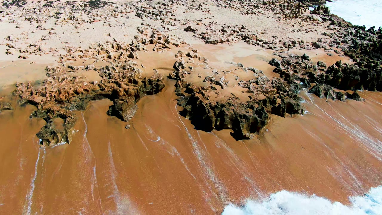 Aerial frame at Punta Buzzards shows white surf pushing into the lower right corner below dark rocks and brown sand, with no visible people.