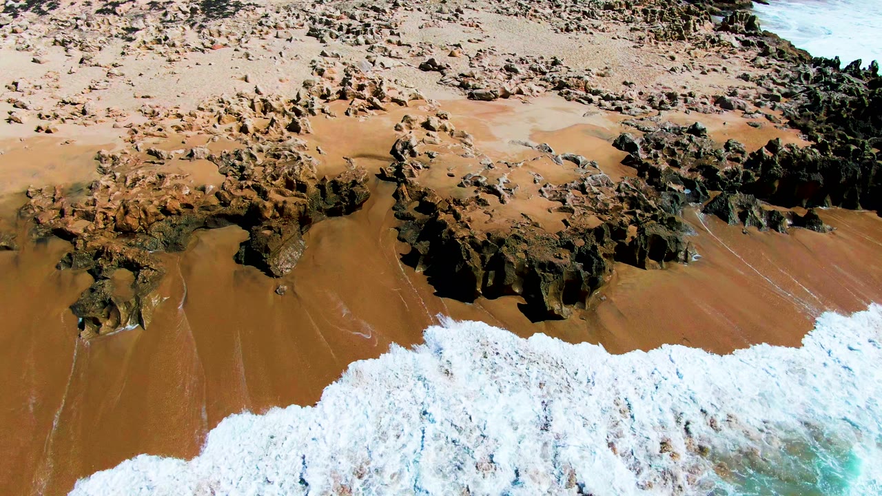 Aerial frame at Punta Buzzards shows a thicker band of white foam crossing the lower beach below jagged rocks and wet brown sand, with no visible people.