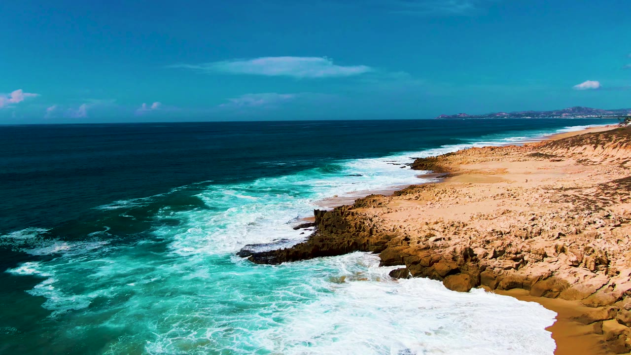 At Punta Buzzards in Baja California Sur, turquoise surf pushes toward dark rocks and tan sand under a blue sky, with no visible people.
