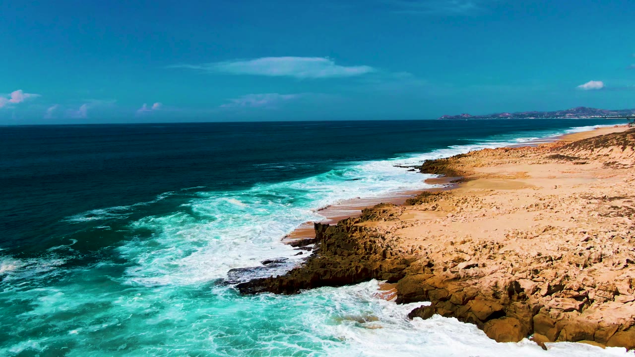 Aerial frame over Punta Buzzards shows white surf widening along the rocky edge while deeper blue water sits offshore, with no visible people.