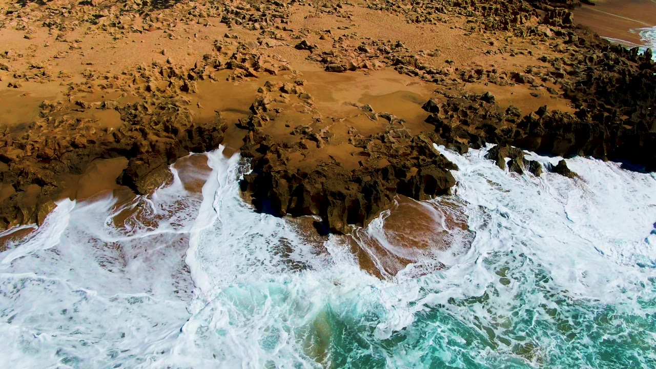 Overhead view at Punta Buzzards, Baja California Sur, with white surf spreading around dark shore rocks onto brown sand and no visible people.