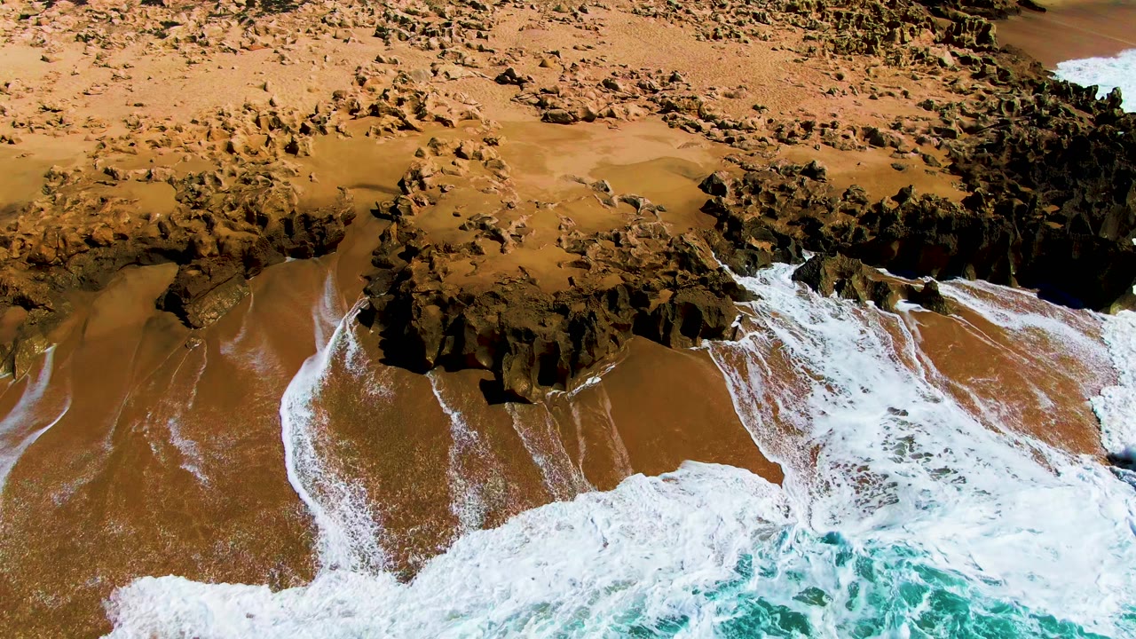 At Punta Buzzards, a broad white wave line presses into the rocky shoreline while wet sand remains visible between foam bands and no people are visible.
