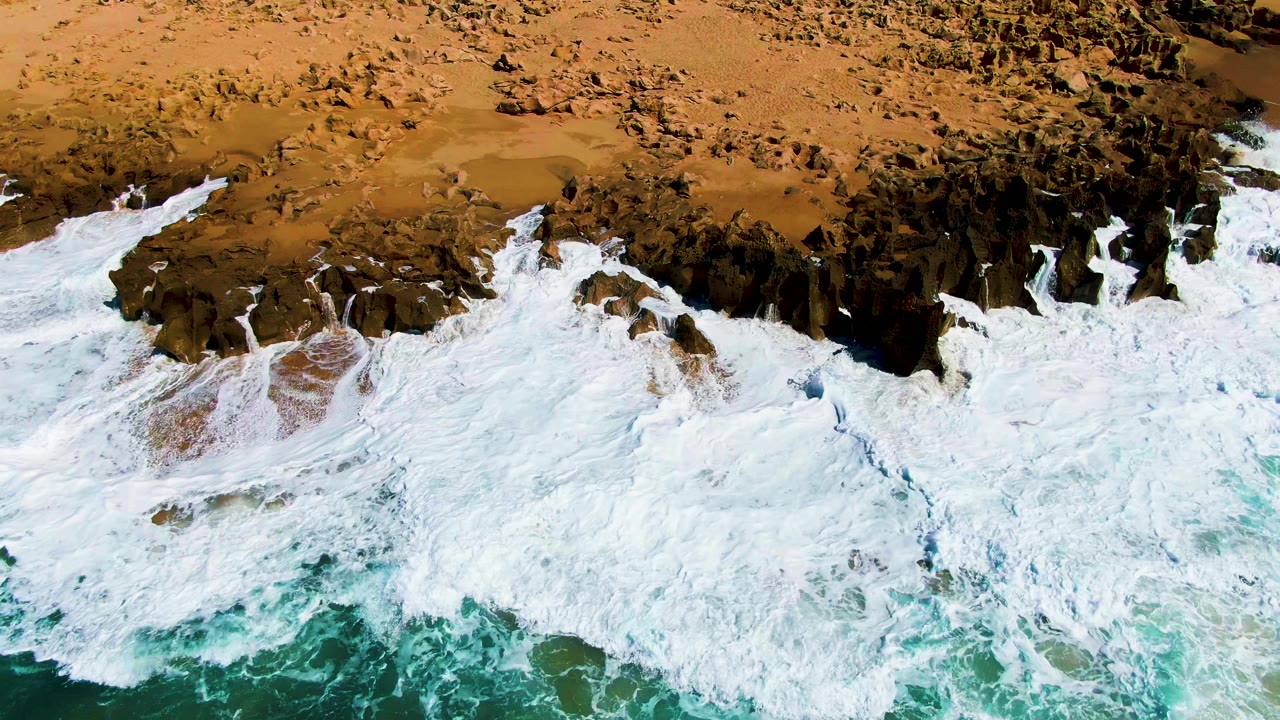 Aerial frame of whitewater pushing farther into jagged shoreline rocks at Punta Buzzards, with dry tan beach across the upper frame and no visible people.