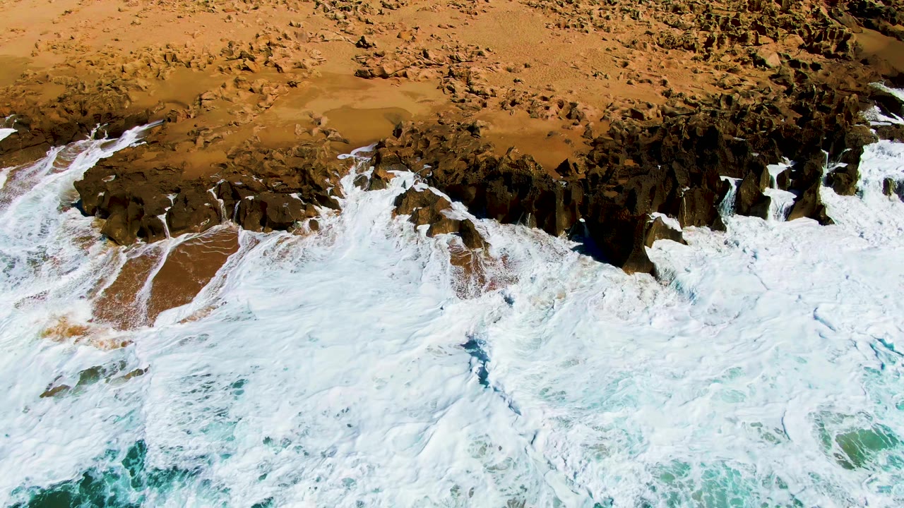 Overhead surf at Punta Buzzards covers much of the rocky beach edge, with streaks of white foam over brown sand and turquoise water at the lower edge, no visible people.