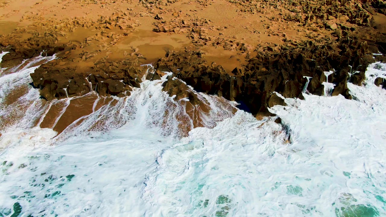 Aerial view at Punta Buzzards as white foam thins into streaks over wet brown sand beside dark rocks, with no visible people on the beach.