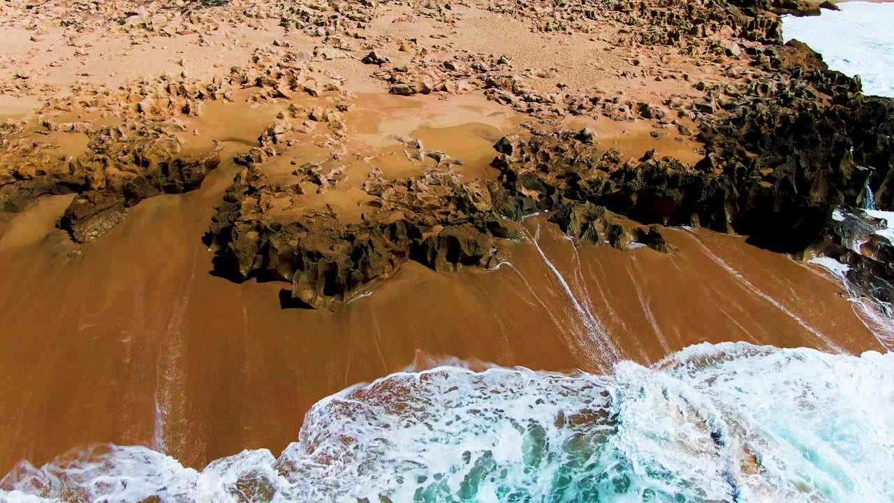Aerial view of surf reaching tan sand below dark coastal rocks.