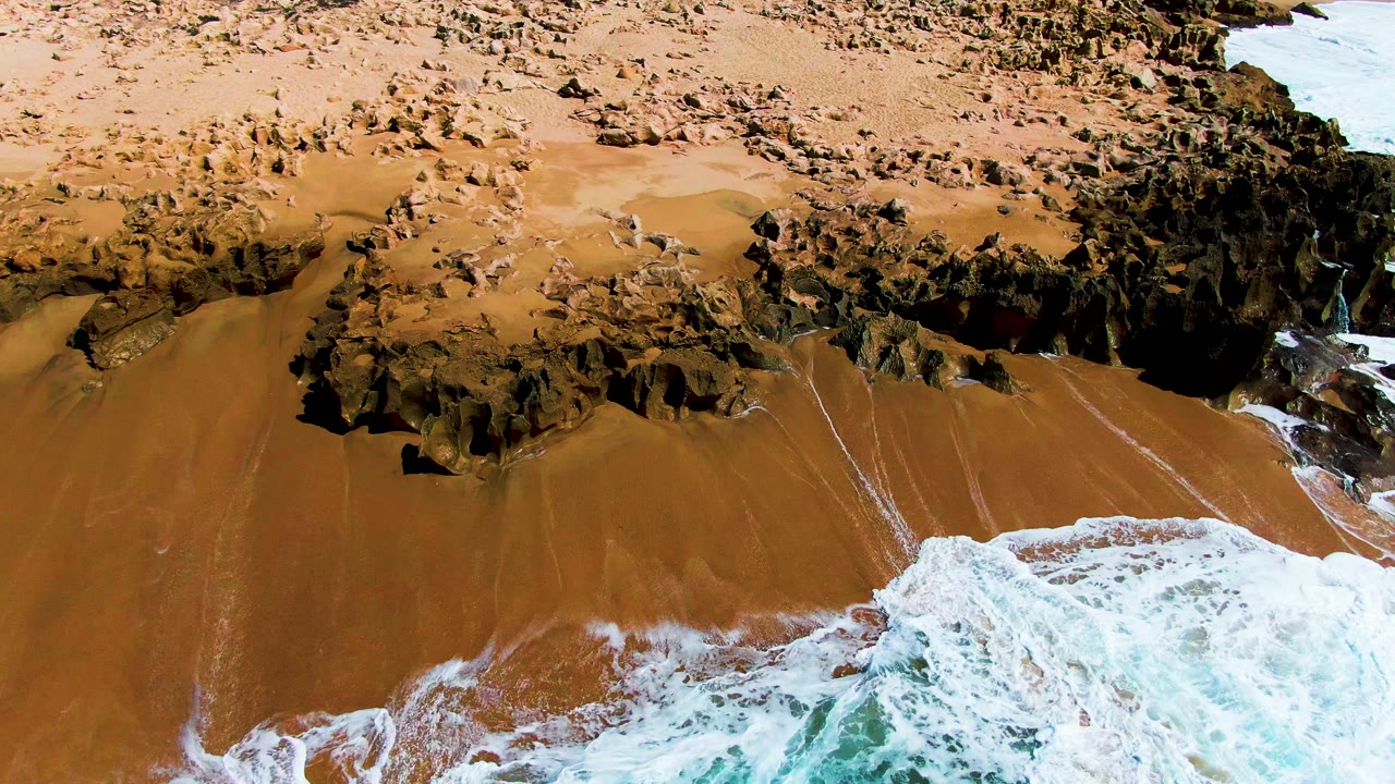 Aerial frame over Buzzards Beach showing white foam advancing across tan sand with dark rock ledges along the upper shoreline, no visible people.