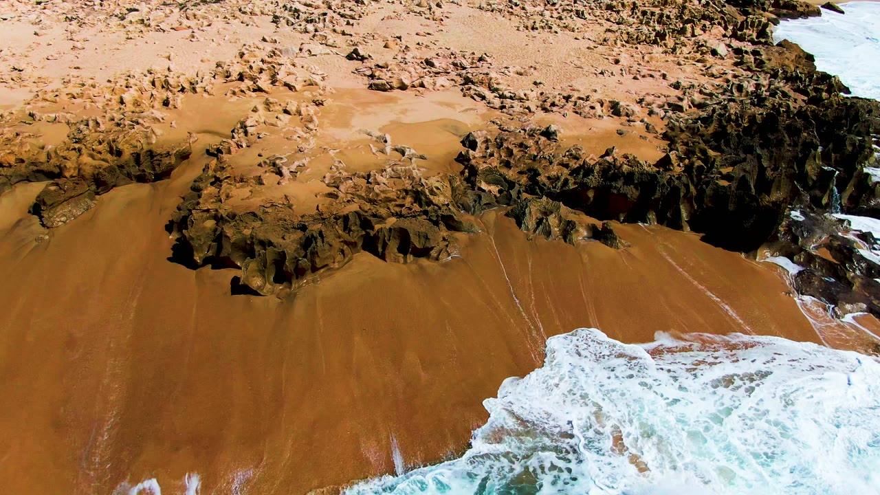 Aerial frame over Buzzards Beach with turquoise water and white foam curling onto the lower right side of the sandy shore beside dark rocks, no visible people.