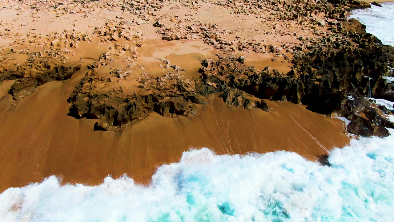Aerial frame over Buzzards Beach where a broad white wave edge pushes up from the bottom of the frame toward tan sand and dark rocky outcrops, no visible people.