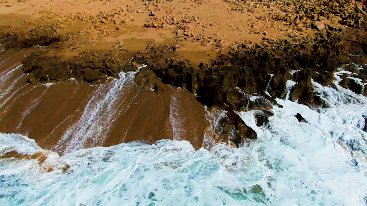 At Buzzards Beach in Baja California Sur, white surf breaks across dark shoreline rocks beside brown sand, with no visible people.