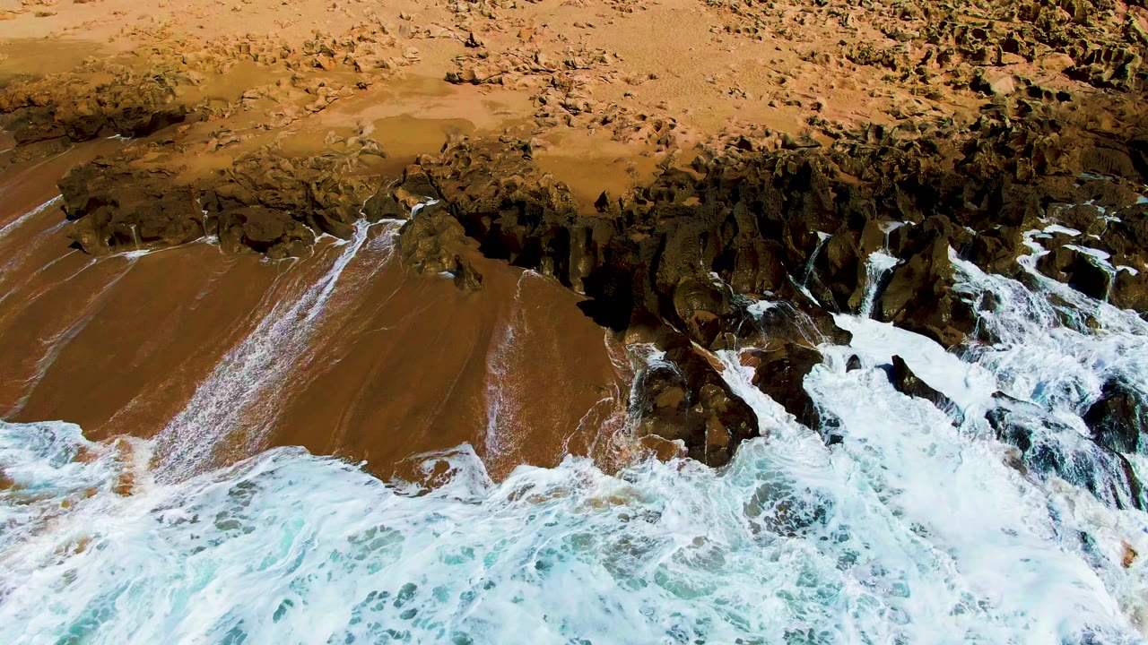 At 0:01, foamy pale blue water advances toward jagged dark rocks and wet brown sand at Buzzards Beach, with no visible people.