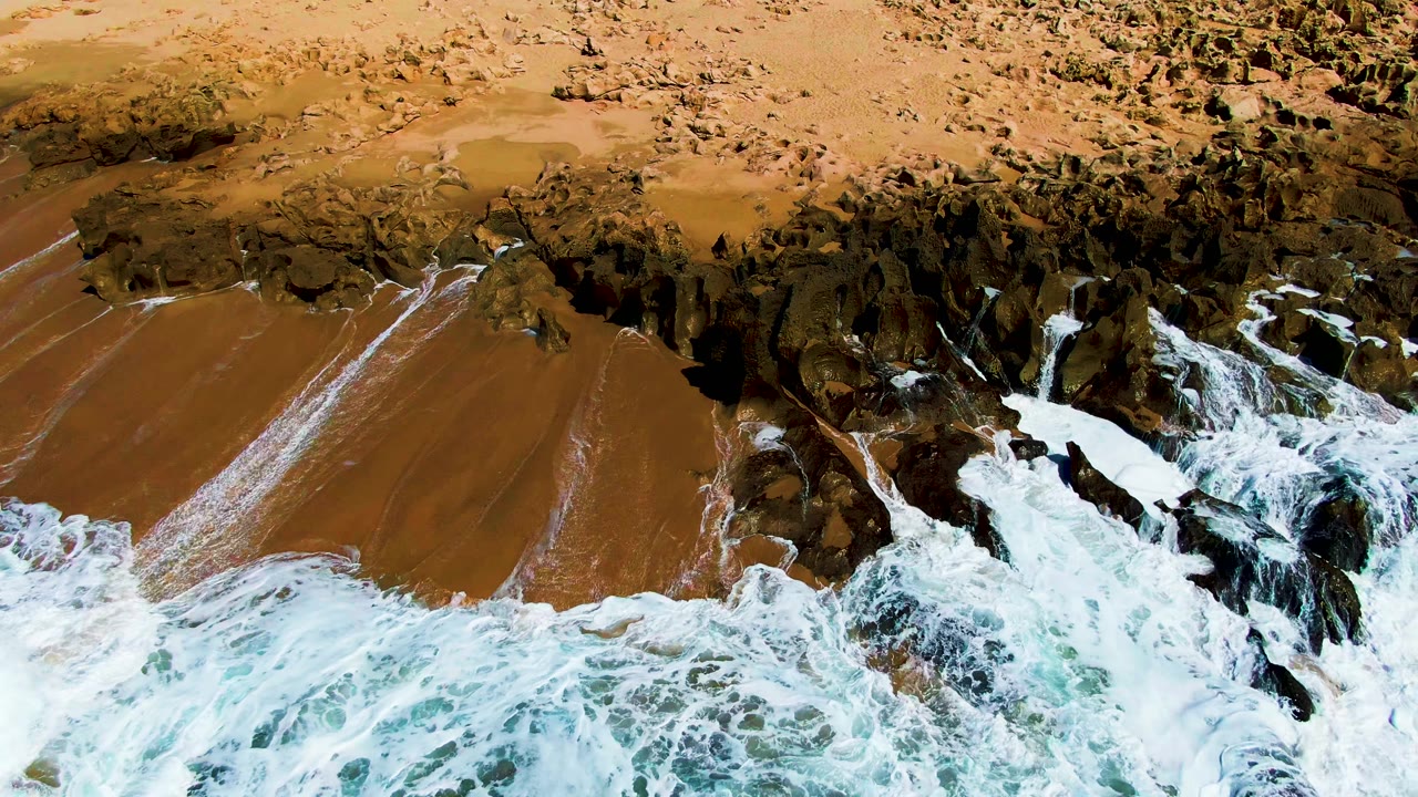 At Buzzards Beach, whitewater churns over the lower frame while dark rock shelves and brown sand remain visible above, with no visible people.
