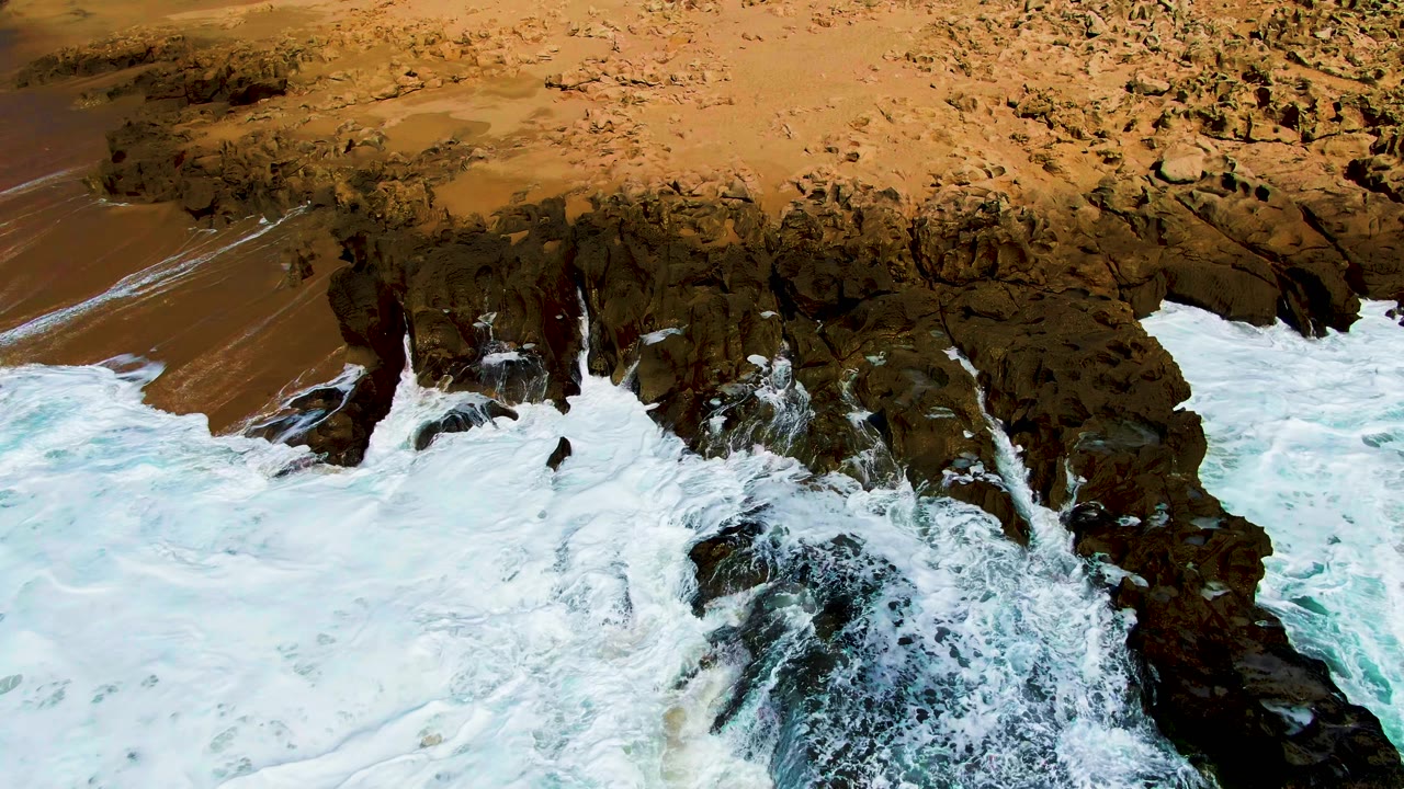 At Punta Buzzards in Baja California Sur, white ocean foam wraps around dark shoreline rocks below a sandy beach, with no visible people.