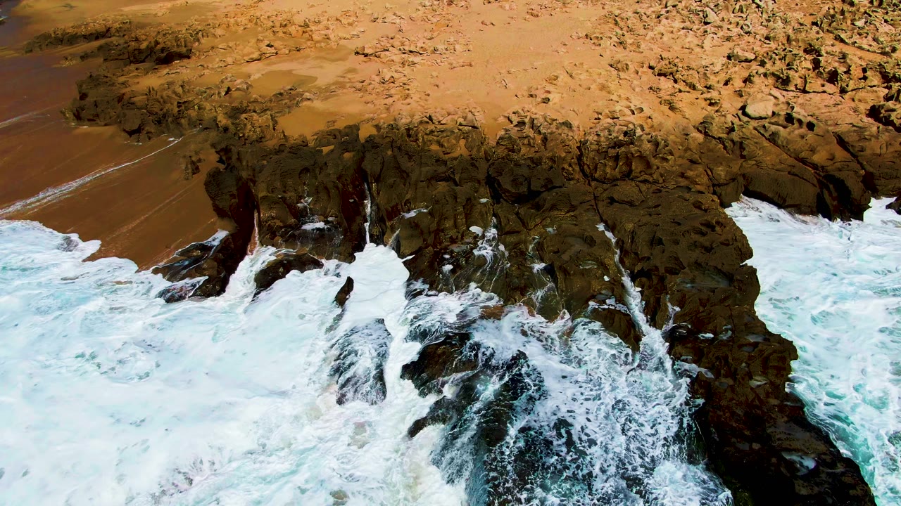 Foamy water presses into the left side of the rocky point while the tan beach remains visible behind it at Punta Buzzards, with no visible people.