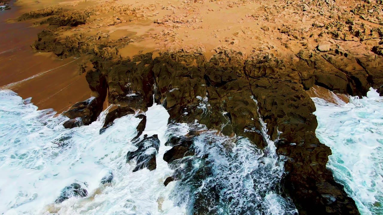 At 0:02, whitewater breaks around jagged dark rocks near the center of the frame as brown sand stretches behind, with no visible people.