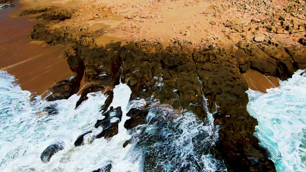 Surf washes along dark rocks beneath an empty sandy beach.