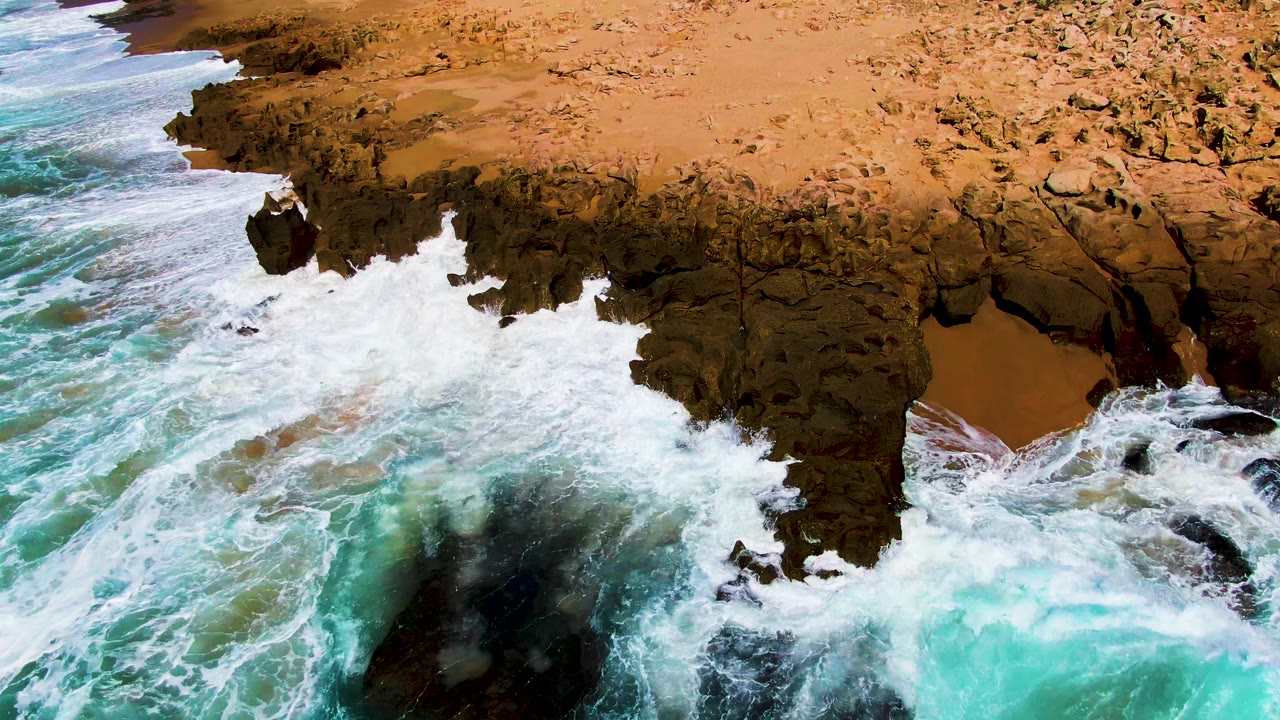 At Punta Buzzards in Baja California Sur, turquoise surf pushes into a dark rocky cliff below tan shoreline terrain, with no visible people.