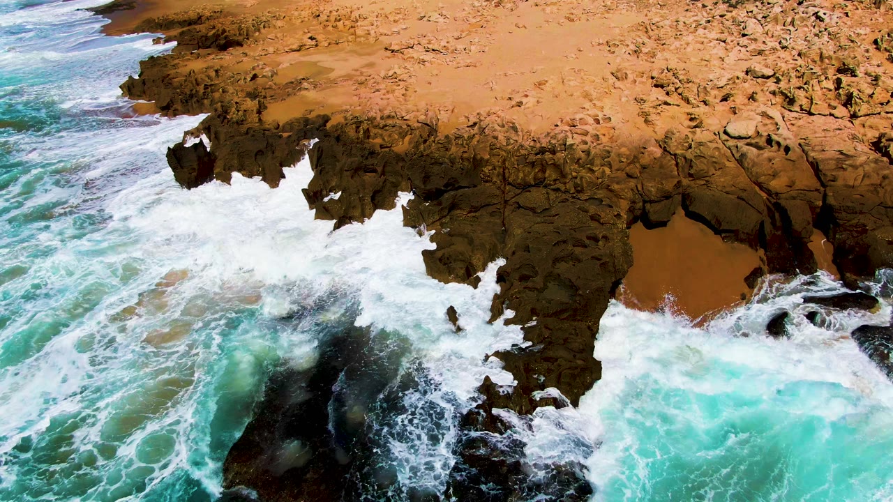At Punta Buzzards in Baja California Sur, white foam spreads across jagged brown rock while turquoise water fills the lower frame, with no visible people.