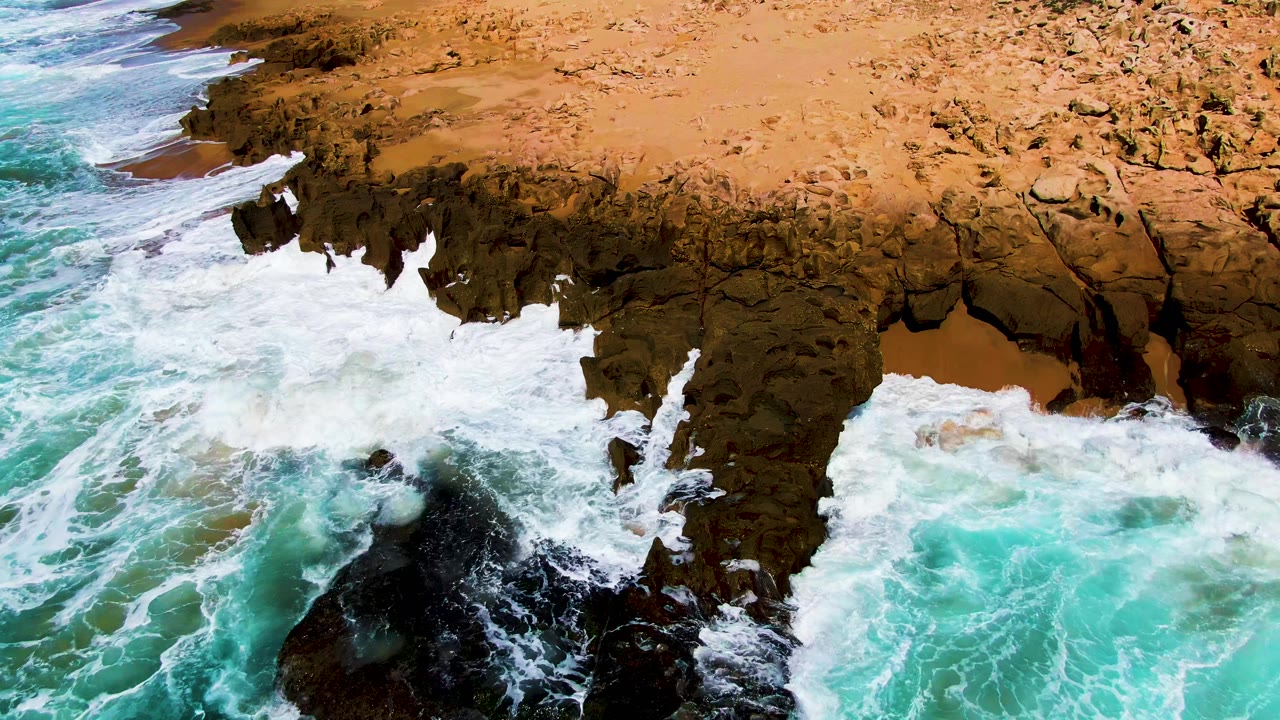 At Punta Buzzards in Baja California Sur, breaking surf wraps around the rocky point and bright turquoise water presses in from the lower right, with no visible people.
