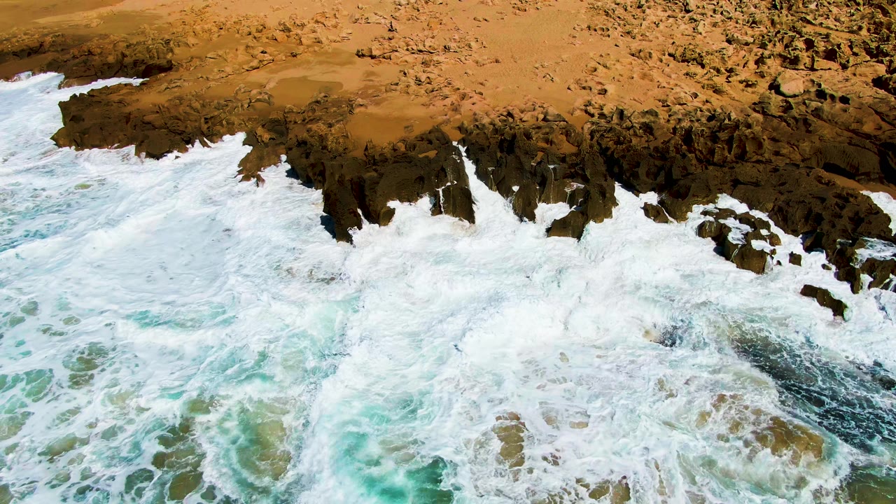 At Punta Buzzards in Baja California Sur, white ocean foam spreads over dark shoreline rocks with tan sand above and no visible people.