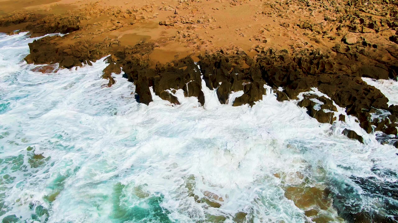 Whitewater pushes farther into jagged dark rock ledges at Punta Buzzards, with turquoise water below, tan shoreline above, and no visible people.