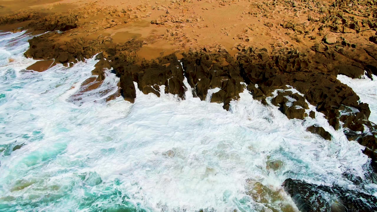 A broad sheet of white surf blankets the lower frame while dark rocks and tan sand define the Punta Buzzards shoreline, with no visible people.