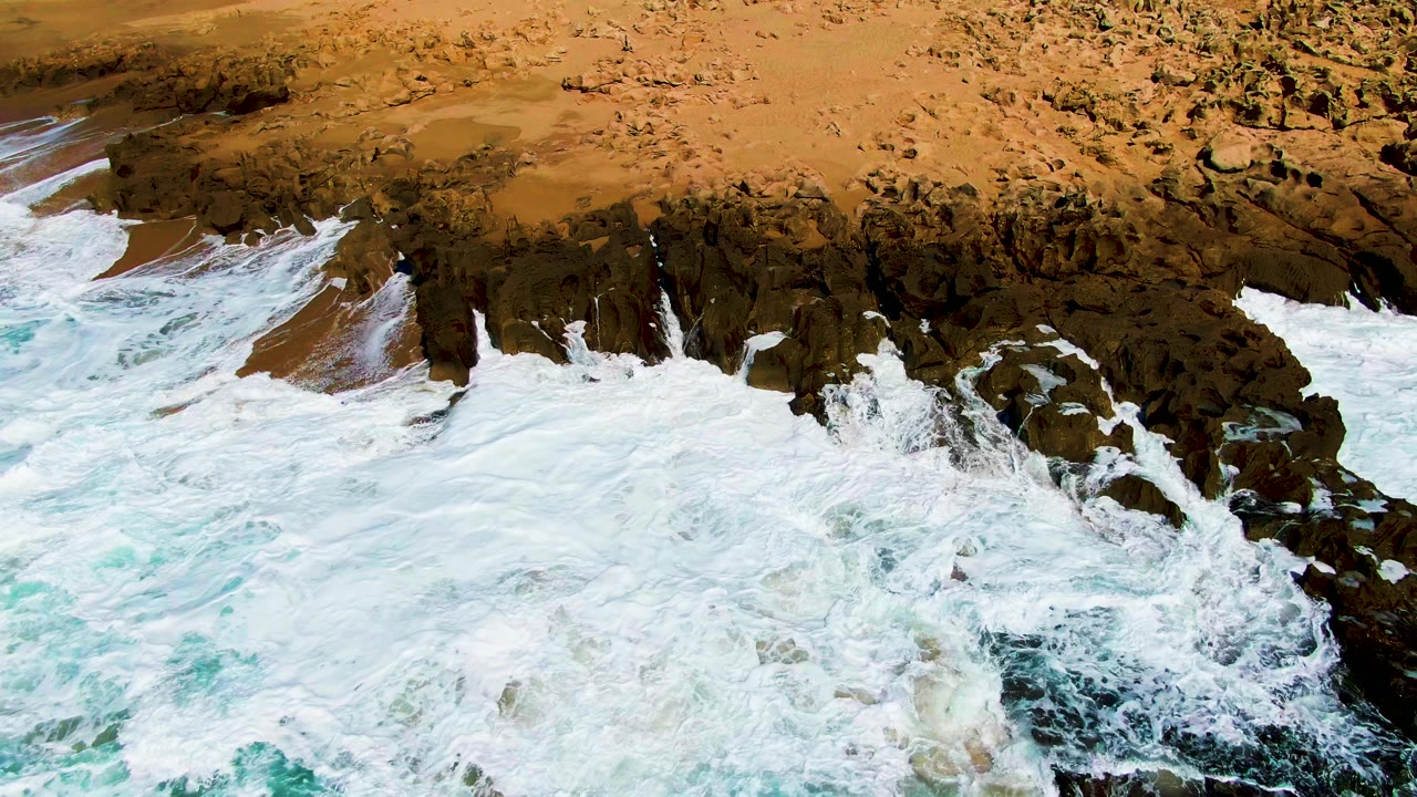 Foam wraps around dark rock outcrops near the sand at Punta Buzzards, with pale blue water at the lower edge and no visible people.