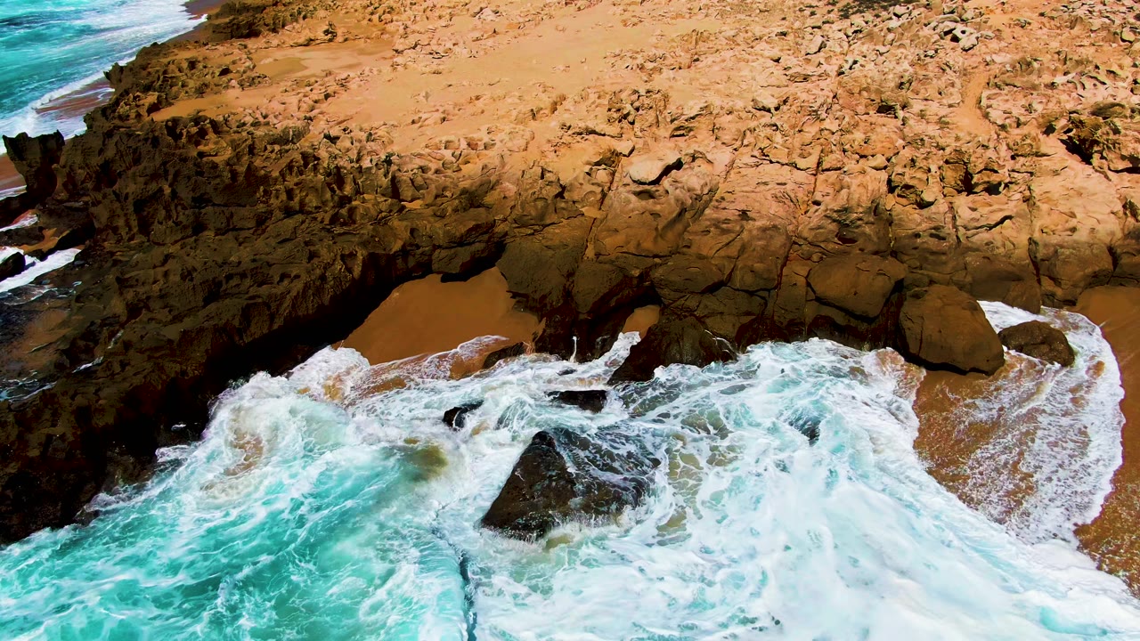 Aerial frame from Punta Buzzards, Baja California Sur, with turquoise surf spreading white foam over dark shoreline rocks beside tan sand and no visible people.