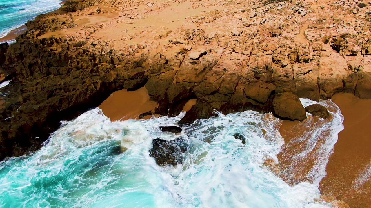At 0:01, white foam wraps around central dark rocks while turquoise water fills the lower left and the dry rocky Punta Buzzards shoreline sits above, with no visible people.