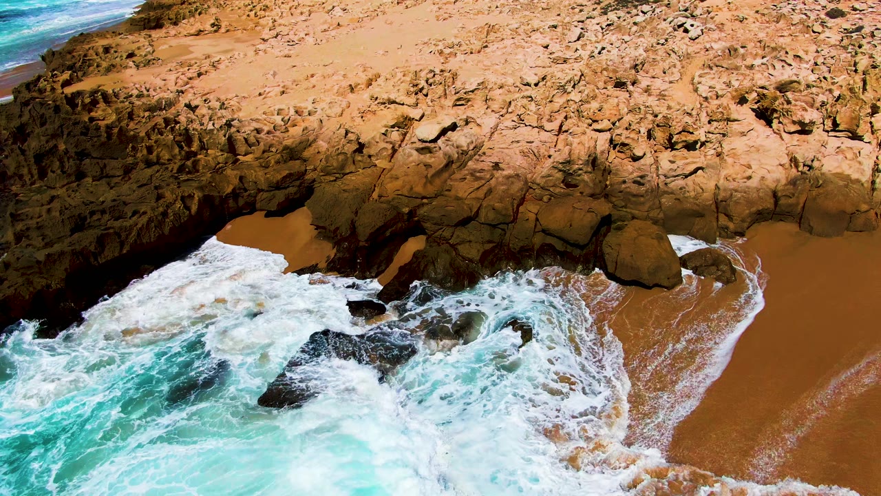 At 0:02, the wave face has flattened into white foam over the rocks, with a broad tan sandy pocket on the right side of the Punta Buzzards beach and no visible people.
