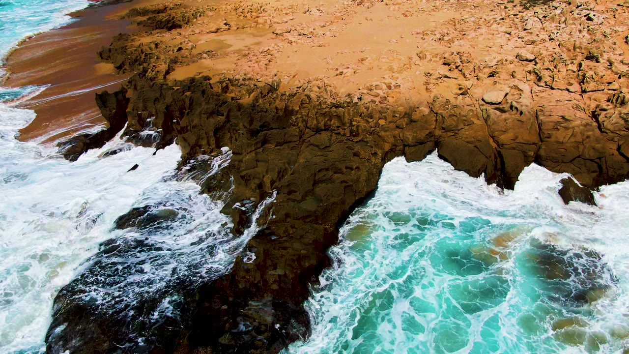 At Punta Buzzards in Baja California Sur, turquoise surf fills the lower frame as waves break beneath a tan rocky coastal arch, with no visible people.
