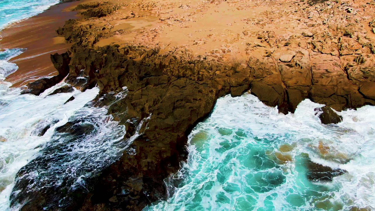 White foam spreads through the dark rock opening at Punta Buzzards while tan shoreline and rough cliff surface sit above, with no visible people.