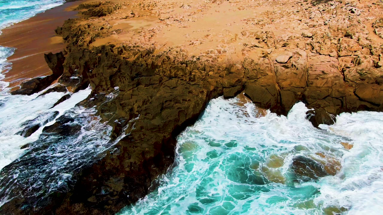 At 0:02, churning turquoise surf presses into the rocky arch at Punta Buzzards as a narrow sandy beach appears along the upper left, with no visible people.