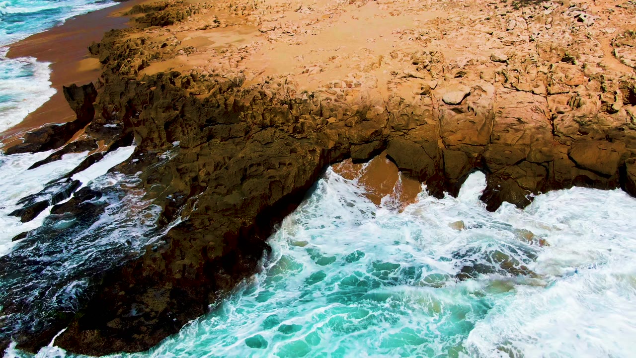 Aerial view of waves breaking against jagged rock below a tan cliff.