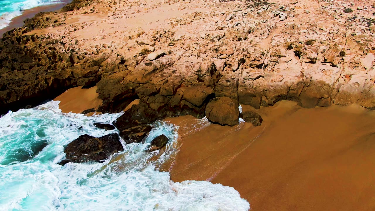 White surf washing around dark rocks beside an orange sandy beach.